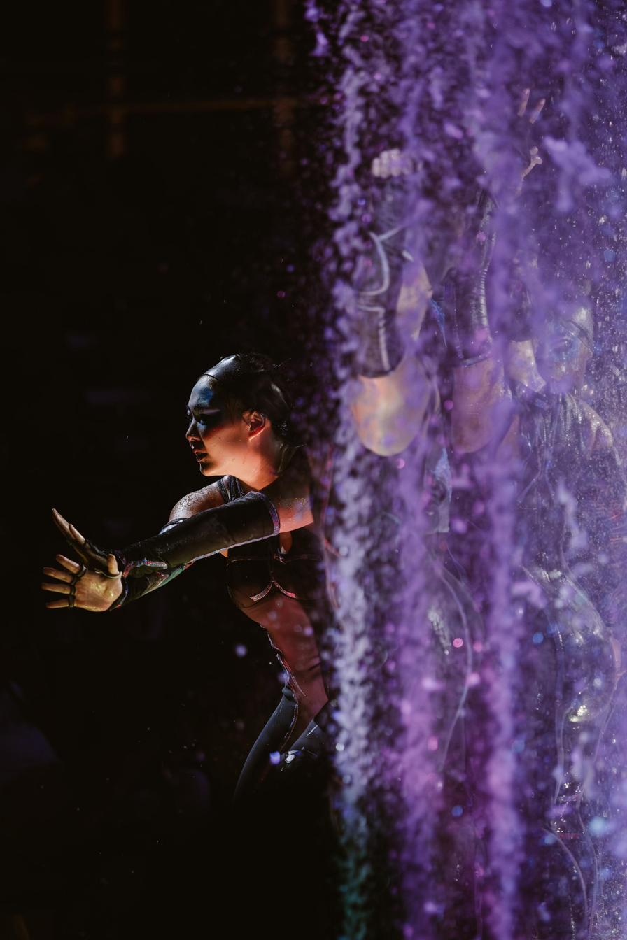 Rebecca reaching through purple-lit water spray on stage